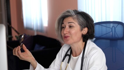 Caucasian doctor with short gray hair wearing white coat, raising hand slightly while holding glasses, smiling gently, deep in thought, expressing composed professionalism. Calm leadership felt.