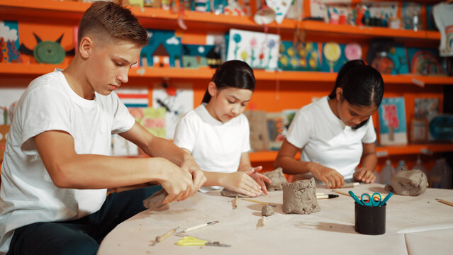 Caucasian smart teenager modeling clay while craving glass of clay at art lesson. Group of multicultural happy student working at dough in pottery workshop. Creative activity concept. Edification.