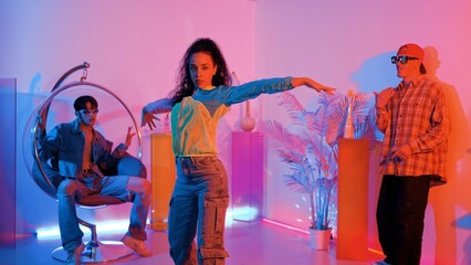 Fototapeta premium Attractive happy dancer looking at camera while smart woman moving at rhyme with neon light. Professional hispanic performer break dancing while wearing colorful cloth with diverse friend. Regalement.