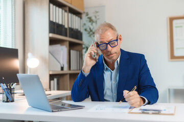 Focused mature businessman talking on mobile phone and reviewing documents while working at office desk