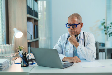 Senior doctor working on laptop in medical office, concentrated mature physician analyzing patient data on computer