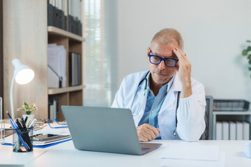 Senior doctor having a headache while using laptop in bright medical office, showing concern and fatigue