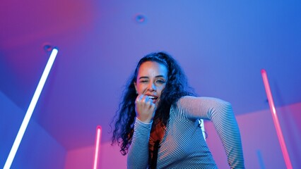 Low angle camera of hispanic dancer looking at camera and dancing to city pop music. Professional attractive hipster moving to hip hop song with purple led or neon light. Stylish cloth. Regalement.