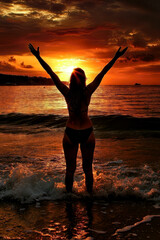 Fototapeta premium A silhouette of an individual with outstretched arms stands in the shallow water, embracing the vibrant sunset over the calm sea while waves lap at their feet.