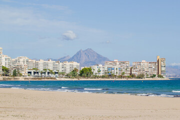 A serene coastal cityscape featuring modern apartment buildings, a sandy beach, and a striking mountain range in the background under a clear blue sky. Alicante, Spain.