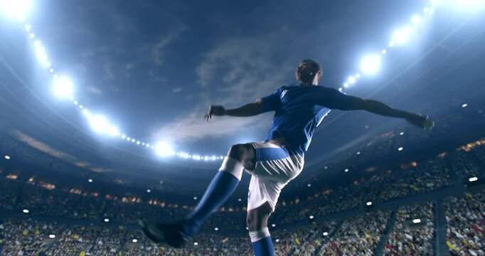 Football player, wearing blue and white uniform, kicks the ball in a jump, in slow motion on the professional football stadium with animated crowd.