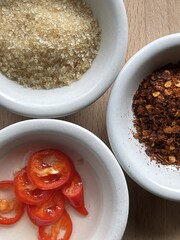 Three small bowls containing sugar, sliced red chili peppers in vinegar, and chili flakes on a wooden surface.
