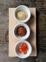 A wooden serving board displays three small bowls containing sugar, chili flakes, and sliced red peppers, set against a rustic background.
