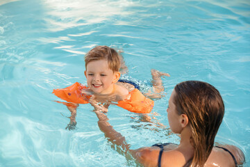 Little cute boy wearing arm floats smiles while learning to swim with his mom in a sunny outdoor...