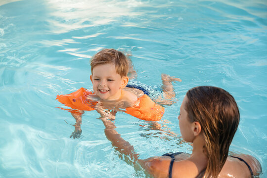 Little cute boy wearing arm floats smiles while learning to swim with his mom in a sunny outdoor swimming pool, enjoying summer fun. 