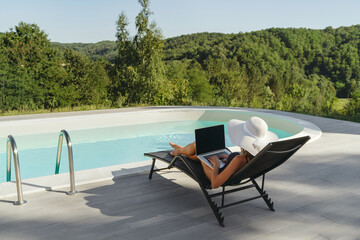 A young woman relaxes on a lounge chair near a pool on sunny day, using her laptop outdoors with a scenic green background. Work-life balance concept. Space for text