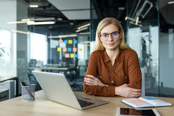 Serious businesswoman sitting at desk with laptop and notebook. Confident mature professional in glasses showing leadership and focus. Perfect for business, corporate themes.