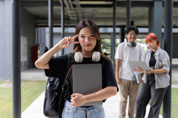 Confident university student gesturing peace sign while holding laptop with friends discussing in the background