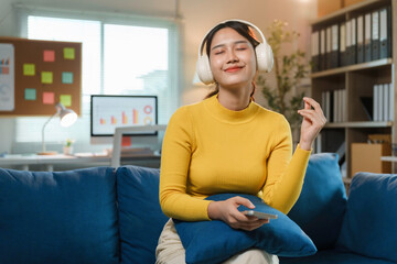 Obraz premium Young asian woman is listening to music with headphones and holding a smartphone while sitting on a sofa in her home office