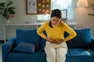 Asian woman suffering from strong abdominal pain while sitting on the sofa at home