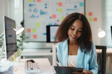 Smiling african american businesswoman taking notes on clipboard analyzing company strategy working at marketing project sitting at desk in startup office