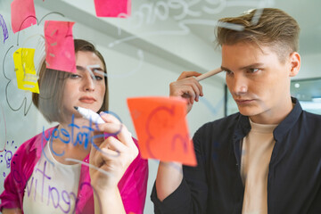 Couple of businesspeople discussing and brainstorming about solving marketing problem in front of glass board at business meeting by using mind map and sticky notes. Working together. Immaculate.