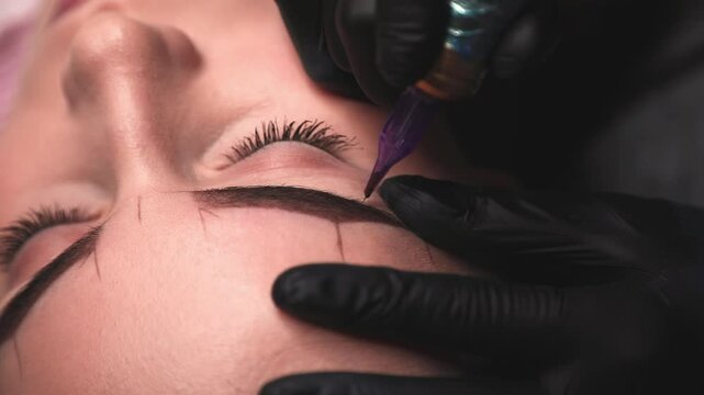 Beautician wearing black gloves uses a tattoo machine to apply permanent makeup on a woman's eyebrow