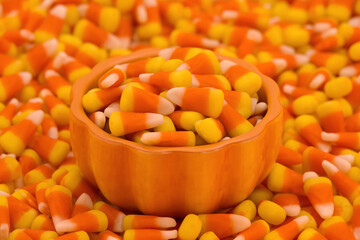 Close up of a small porcelain pumpkin bowl filled with candy corn spilling over onto a background of the yellow orange and white striped traditional Halloween candy.
