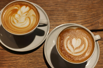 Overhead view of two white cups of cappuccino with heart and leaf latte art  on a rustic wood table in a coffee shop, capturing cafe ambiance and coffee culture. Copy space
