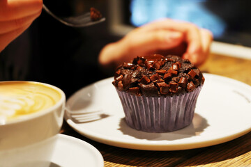 Chocolate muffin with chocolate chunks on a plate beside a latteon the table in coffee shop. Young womans hand holding a fork ready to taste delicious dessert. Selective focus