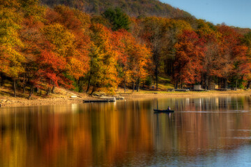 A serene lake scene captures vivid autumn colors as the trees reflect on the calm water. A lone boat drifts gently, enhancing the tranquil atmosphere.