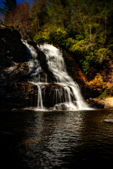 Obraz premium Water cascades down a rocky cliff into a tranquil pool, surrounded by lush greenery and trees. Sunlight highlights the vibrant colors of the foliage, creating a serene and natural scene.