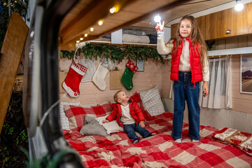 Fototapeta premium Young girl and boy sibling enjoy holiday time in a cozy camper, decorated with festive lights, stockings, and Christmas décor. Warm family moments in a mobile home.