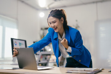 Asian businesswoman in a blue suit is raising her fist in excitement while looking at her laptop in...