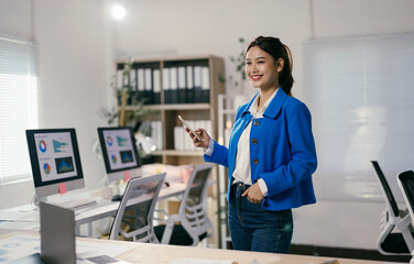 Fototapeta premium Asian businesswoman smiling in a modern office with a smartphone and computer screens showing charts, reflecting a tech-driven work environment