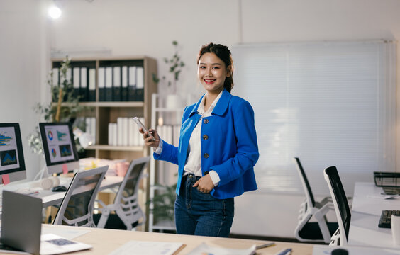 Young asian businesswoman is smiling and holding a smartphone while standing in a modern office with computers and documents on desks