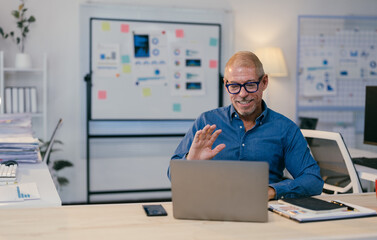 Happy mature manager greeting colleagues during online meeting, waving hand at laptop screen in modern office, sitting at desk with financial documents and charts