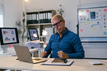 Senior businessman sitting at his desk, clenching his fists and analyzing charts on his laptop,...
