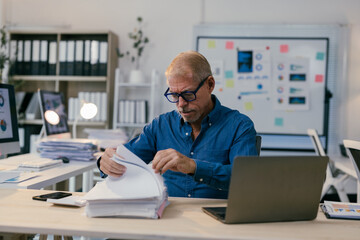Focused mature manager reviewing paperwork, surrounded by office supplies and technology, diligently working on a project
