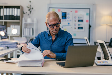 Focused mature professional reviewing paperwork and using a laptop, managing tasks and projects efficiently in a busy office environment with charts and graphs displayed on a whiteboard