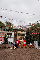 Woman in cozy plaid blanket sits outside a holiday-decorated camper. Christmas tree, guitar, and rustic holiday decor create a warm and inviting festive atmosphere.