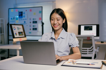 Young happy business woman using laptop computer, working with documents and diagrams, smiling and sitting at her desk in modern office at night