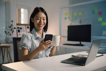 Young asian businesswoman smiling and using smartphone while drinking coffee, sitting at her desk in a modern office with laptop and computer monitor in background