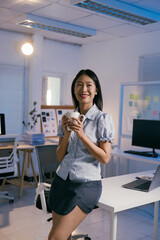 Happy businesswoman holding a cup of coffee and smiling while leaning on a desk in a modern office, enjoying a relaxing break during her workday