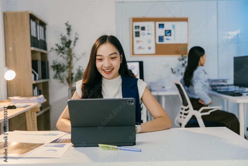 Poster Young office worker using tablet and smiling while working at her desk in a bright and modern office, with a colleague working in the background