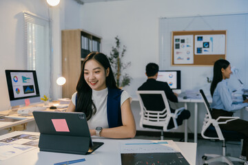Young businesswoman using tablet and smiling while working in modern office with colleagues in background, focusing on concept of productivity and teamwork in business environment