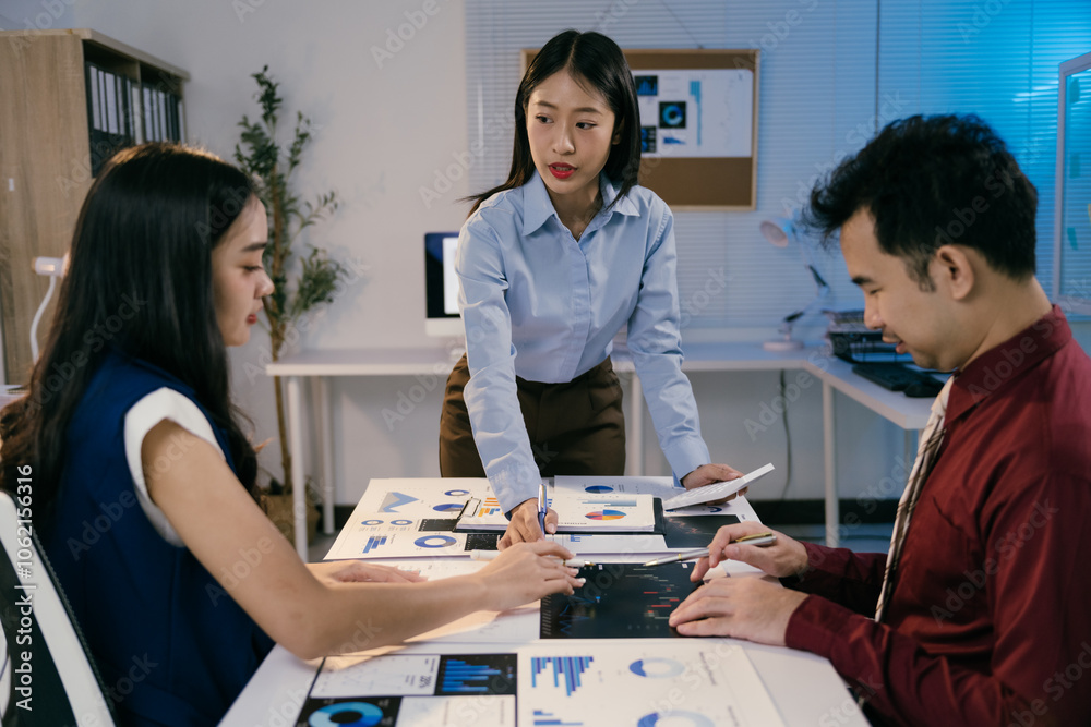 Poster Three asian businesspeople are working late in a brightly lit modern office, collaborating on data analysis using printed charts and reports, demonstrating teamwork and dedication