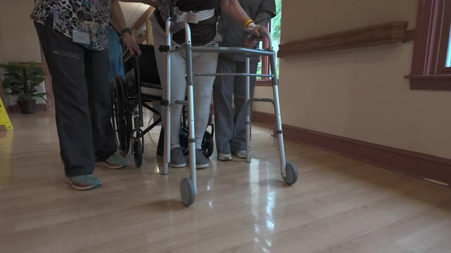 A patient using a walker is assisted by a caregiver, while a wheelchair is nearby in a hospital hallway with wood flooring.