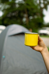 Young woman hand holding a yellow coffee cup while outdoors on the camping site with a tent and a tree blurred in the background.