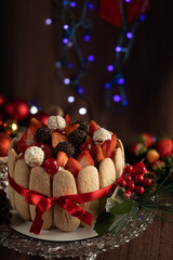 Festive Christmas Charlotte cake adorned with strawberries, cherries, chocolate truffles, and whipped cream, surrounded by ladyfingers and a red ribbon, evoking holiday cheer and winter celebrations.
