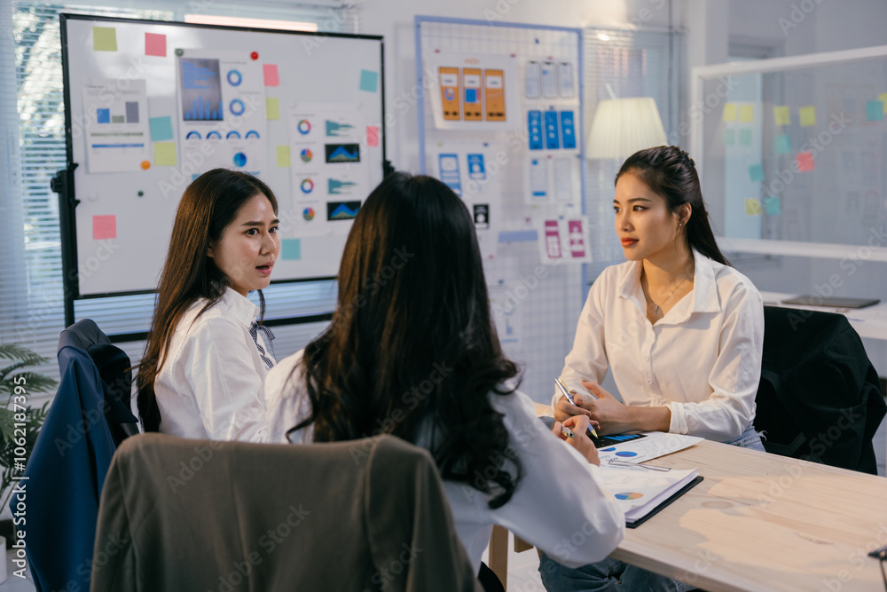 Poster three asian businesswomen are sitting at a table in a modern office, actively discussing charts and 