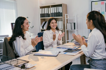 Three asian businesswomen are sitting at a desk, clapping their hands and smiling, celebrating the success of their project after a productive meeting in a modern office