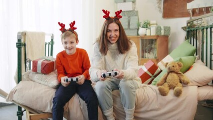 A joyful mother and son play video games together with joysticks, celebrating the Christmas holiday with festive antlers and gifts around them, and son losing
