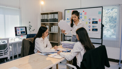 Three asian businesswomen are working together in a modern office, analyzing financial reports, charts, and graphs while discussing company performance and strategies © PaeGAG