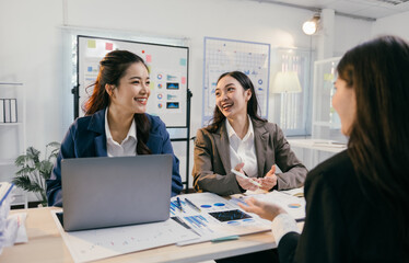 Three asian businesswomen are sitting at a desk in a modern office, smiling and discussing charts and data, working together on a laptop